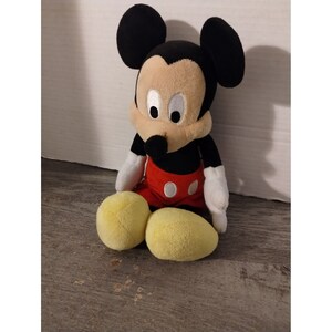 May include: A plush Mickey Mouse toy sitting with his legs crossed. He is wearing red shorts with white polka dots and has black ears and a black nose.