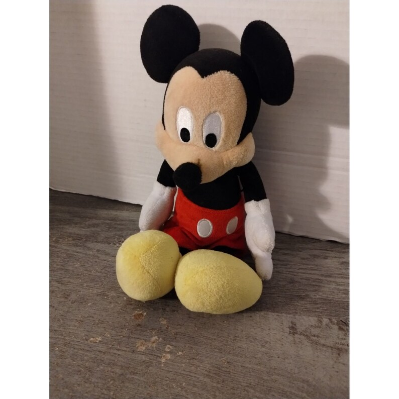 May include: A plush Mickey Mouse toy sitting with his legs crossed. He is wearing red shorts with white polka dots and has black ears and a black nose.