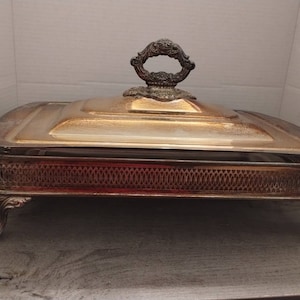May include: Silver serving dish with a glass lid and ornate handle. The dish has a decorative, patterned base and four feet.