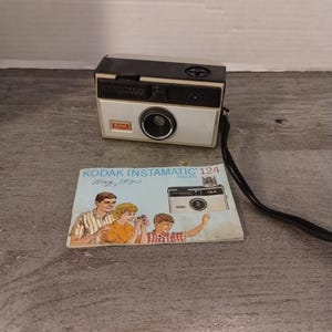 May include: A vintage Kodak Instamatic 124 camera with a black strap and a white and black body. The camera has a red Kodak logo and the text "Instamatic" and "124" printed on it. The camera is accompanied by a vintage instruction manual with the text "Kodak Instamatic 124 Camera May, 1970".