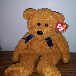 May include: A brown teddy bear with a blue ribbon around its neck. The bear has a red nose and black eyes. The bear is sitting on a wooden surface. The bear has a Ty tag attached to its ear.
