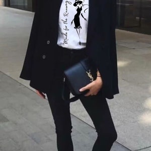 May include: A woman wearing a white t-shirt with the text "Made for the Runway" and a black silhouette of a woman in a dress. She is also wearing a black blazer, black jeans, and white sneakers.