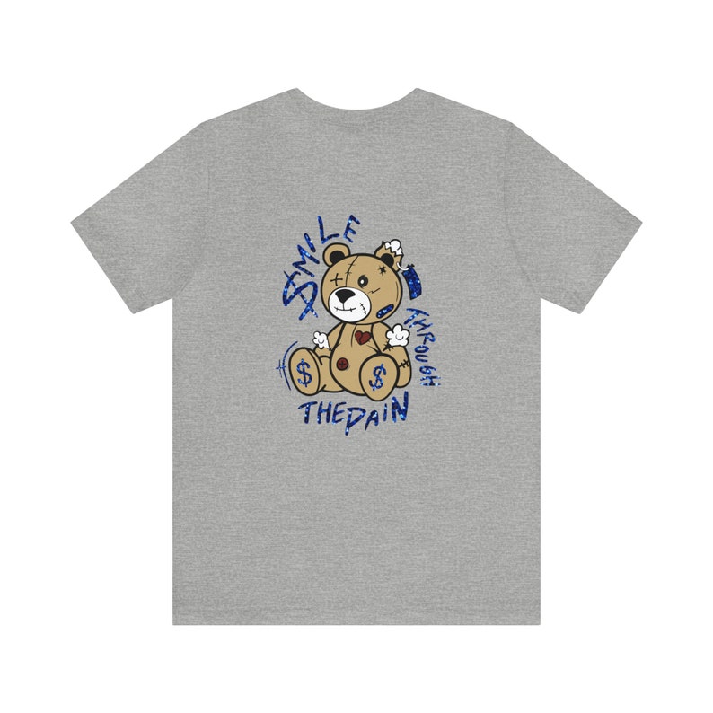 Puede incluir: Camiseta gris con un gr&aacute;fico de un oso de peluche de dibujos animados. El oso es marr&oacute;n y tiene puntos de sutura en su cuerpo. El texto "Smile Through The Pain" est&aacute; escrito en azul alrededor del oso.