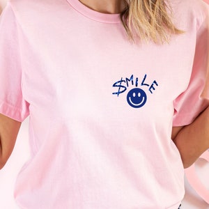 Puede incluir: Una camiseta rosa con un gr&aacute;fico azul que dice "SMILE" con una carita sonriente azul.
