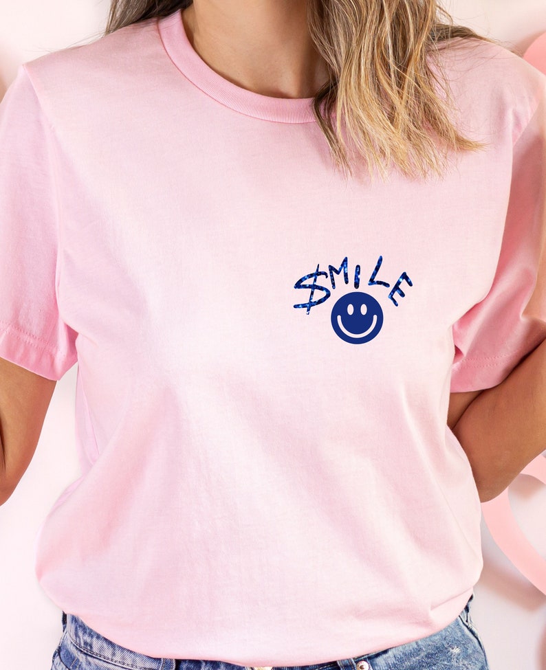 Puede incluir: Una camiseta rosa con un gr&aacute;fico azul que dice "SMILE" con una carita sonriente azul.