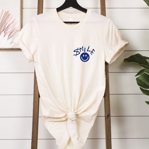 Puede incluir: Una camiseta de color crema con un gr&aacute;fico azul de una cara sonriente y la palabra "SMILE" en la parte delantera.