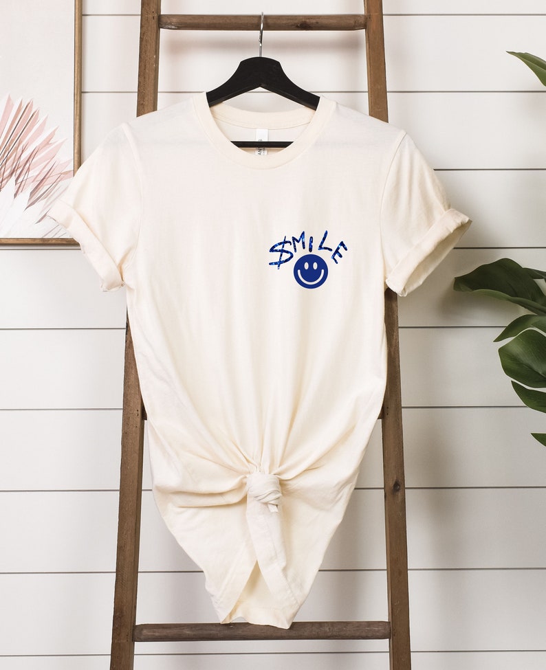 Puede incluir: Una camiseta de color crema con un gr&aacute;fico azul de una cara sonriente y la palabra "SMILE" en la parte delantera.