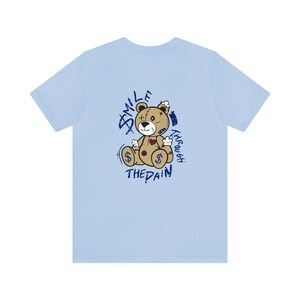 Puede incluir: Camiseta azul claro con un gr&aacute;fico de un oso de peluche desgastado. El gr&aacute;fico dice "Smile Through The Pain" en letras de purpurina azul.