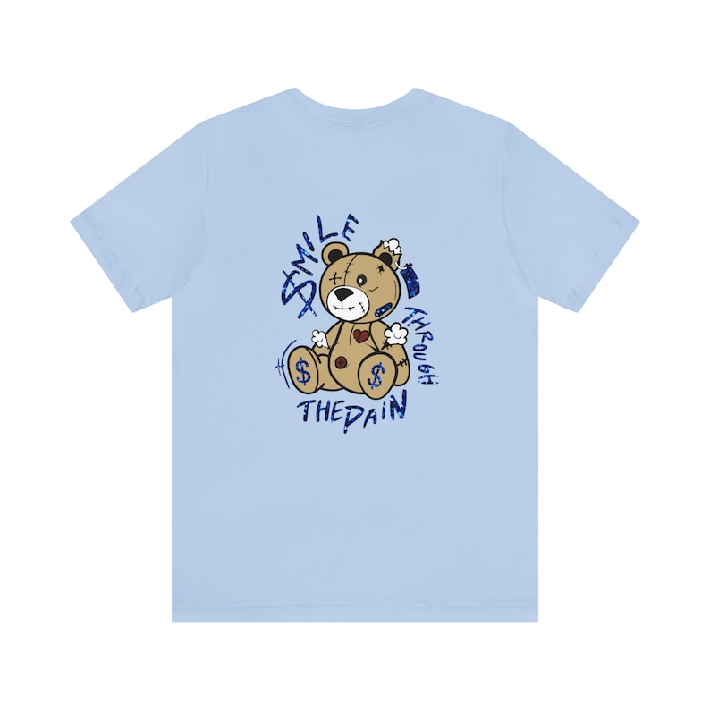 Puede incluir: Camiseta azul claro con un gr&aacute;fico de un oso de peluche desgastado. El gr&aacute;fico dice "Smile Through The Pain" en letras de purpurina azul.