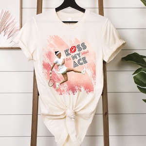 以下が含まれることがあります： テニス選手のグラフィックと「Kiss Ons My Ace」の文字が入ったクリーム色のTシャツ。デザインには赤いキスマークとピンクとピーチの背景が含まれています。Tシャツは黒いハンガーに掛けられています。