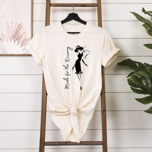 May include: A cream-colored t-shirt with a black silhouette of a woman wearing a hat and dress. The text "Made for the Runway" is printed vertically on the left side of the shirt.