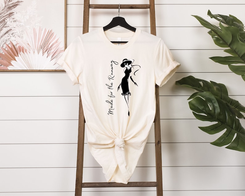 May include: A cream-colored t-shirt with a black silhouette of a woman wearing a hat and dress. The text "Made for the Runway" is printed vertically on the left side of the shirt.