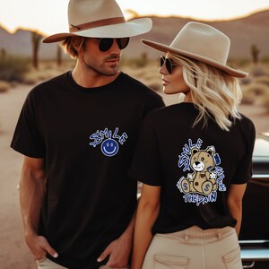 Puede incluir: Dos personas con camisetas negras con dise&ntilde;os gr&aacute;ficos azules. El hombre lleva un sombrero vaquero marr&oacute;n y gafas de sol. La mujer lleva un fedora marr&oacute;n y gafas de sol. La camiseta del hombre dice "Smile at the pain" con una cara sonriente. La camiseta de la mujer dice "Smile through the pain" con un gr&aacute;fico de un osito de peluche.
