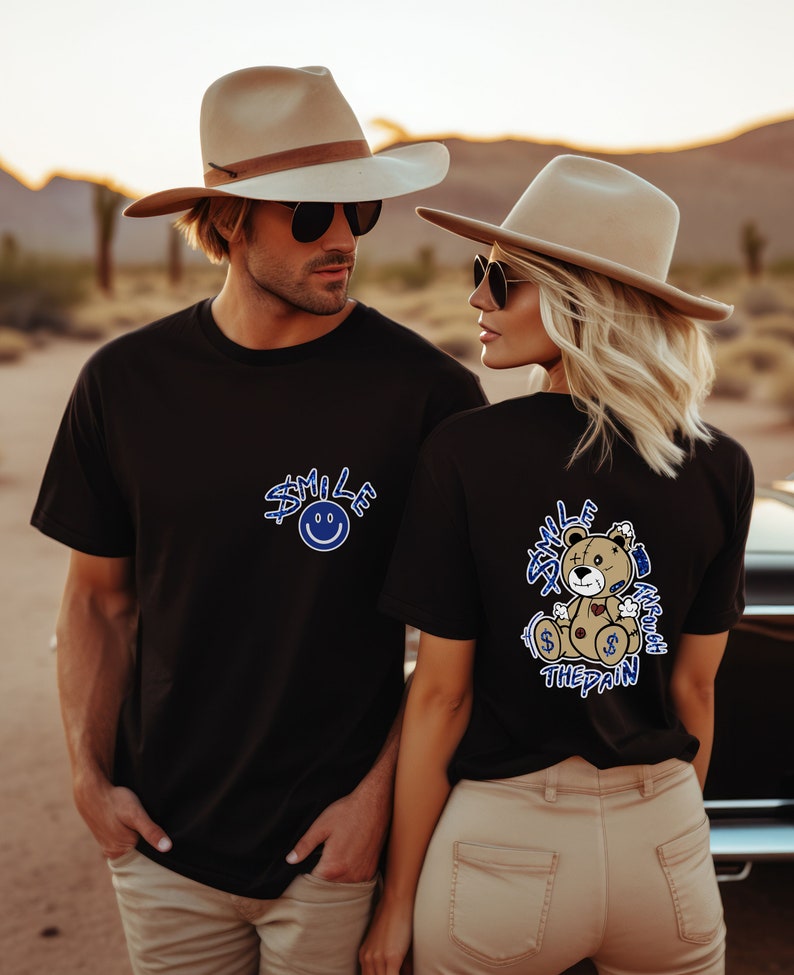 Puede incluir: Dos personas con camisetas negras con dise&ntilde;os gr&aacute;ficos azules. El hombre lleva un sombrero vaquero marr&oacute;n y gafas de sol. La mujer lleva un fedora marr&oacute;n y gafas de sol. La camiseta del hombre dice "Smile at the pain" con una cara sonriente. La camiseta de la mujer dice "Smile through the pain" con un gr&aacute;fico de un osito de peluche.