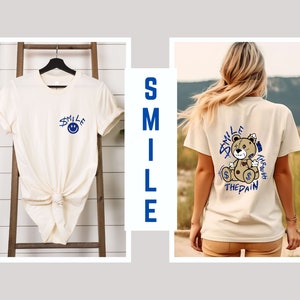 Puede incluir: Dos camisetas blancas con dise&ntilde;os diferentes. La primera camiseta tiene una cara sonriente azul con la palabra "smile" escrita encima. La segunda camiseta tiene un oso de dibujos animados con las palabras "Smile Through The Pain" escritas alrededor.