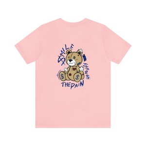 Puede incluir: Camiseta rosa con un gr&aacute;fico de un oso de peluche desgastado con el texto "Smile Through The Pain" en purpurina azul.