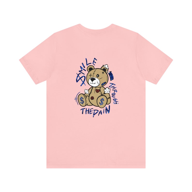 Puede incluir: Camiseta rosa con un gr&aacute;fico de un oso de peluche desgastado con el texto "Smile Through The Pain" en purpurina azul.
