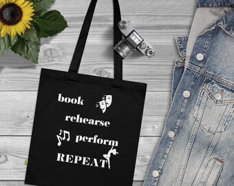 May include: Black tote bag with white text that reads "book rehearse perform repeat". The bag also features a white musical note, two white theater masks, and a white silhouette of a dancer.