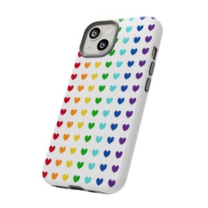 Puede incluir: Una funda de teléfono blanca con un patrón de corazones arcoíris. La funda tiene un recorte para el objetivo de la cámara.