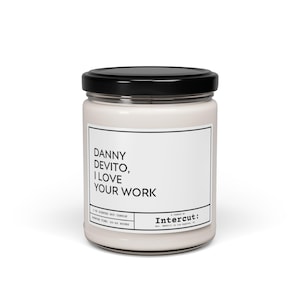 May include: A white glass candle jar with a black lid. The label reads "Danny Devito, I Love Your Work". The candle is made by Intercut.