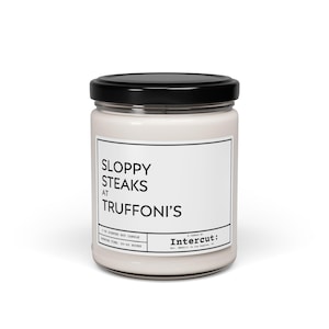Può includere: Una candela in vetro bianco in un barattolo con un coperchio nero. L'etichetta recita "Sloppy Steaks at Truffoni's". La candela è realizzata da Intercut.