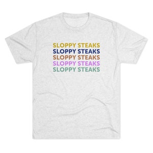 Può includere: Una t-shirt color grigio melange con il testo "SLOPPY STEAKS" ripetuto cinque volte in diversi colori: oro, blu navy, marrone, viola e verde acqua.