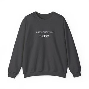 May include: Dark gray sweatshirt with the text "PREVIOUSLY ON THE OC" in white. The long-sleeved crew neck sweatshirt is a solid color and has a simple, classic design. The text is centered on the chest.