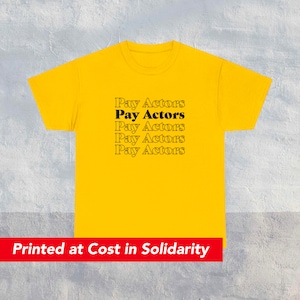 Puede incluir: Camiseta amarilla con el texto "Pay Actors" repetido cinco veces en negro. El texto está impreso en negrita, fuente sin serifas. La camiseta tiene mangas cortas y cuello redondo. El texto "Printed at Cost in Solidarity" está impreso en rojo en la parte inferior de la imagen.