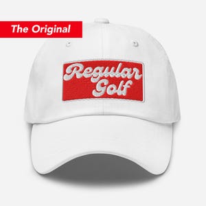 Op de afbeelding: Witte baseballpet met een rode rechthoekige patch met de woorden "Regular Golf" in witte letters geborduurd. De pet heeft een gebogen klep en een gestructureerde kroon. De tekst "The Original" staat in een rode rechthoek.