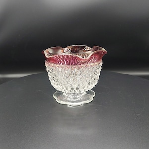 May include: A clear glass dish with a ruffled, ruby red rim and a diamond-cut pattern on the bowl. The dish sits on a clear, scalloped base, showcasing a vintage aesthetic. The bowl is likely used for serving small portions.