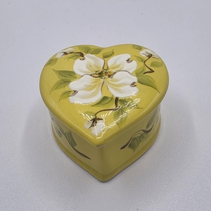 May include: A heart-shaped, yellow trinket box with a white floral design on the lid. The box features a glossy finish and is decorated with green leaves and brown stems. A decorative storage item.