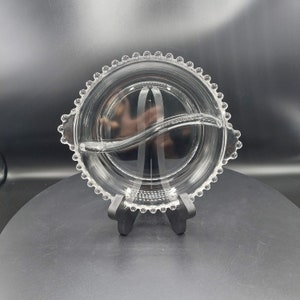 May include: A clear glass divided serving dish with a beaded rim. The dish has two sections separated by a curved divider.