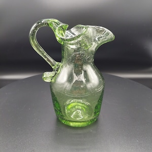 May include: A small, green glass pitcher with a ruffled lip and a handle. The pitcher has a textured surface and is clear.
