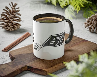 May include: White ceramic mug with a black rim and handle. The mug has a black and white graphic of the number 65 with a signature underneath. The mug is filled with coffee and steam is rising from the top.