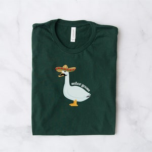 May include: A dark green t-shirt featuring a white goose wearing a sombrero and a moustache. The text "senor goose" is printed below the goose.
