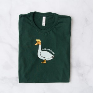 May include: Dark green t-shirt with a cartoon goose illustration. The goose has a yellow beak and a blonde hairstyle, with the words "Indicted Goose" in white text. The shirt is folded neatly on a white marble surface.