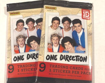 May include: Two unopened One Direction trading card packs. Each pack features a photo of the band members and the text "ONE DIRECTION". The packs also state "TRADING CARDS PLUS 1 STICKER PER PACK".