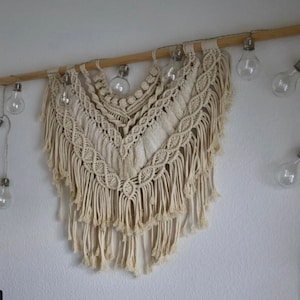 Pode incluir: Uma tape&ccedil;aria de macram&eacute; branca com uma haste de madeira na parte superior. A tape&ccedil;aria tem um design de franjas e &eacute; decorada com uma corda de luzes de vidro transparente.