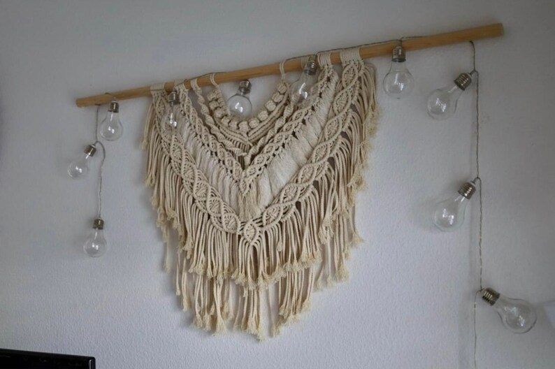 Pode incluir: Uma tape&ccedil;aria de macram&eacute; branca com uma haste de madeira na parte superior. A tape&ccedil;aria tem um design de franjas e &eacute; decorada com uma corda de luzes de vidro transparente.