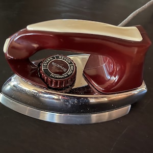 Puede incluir: Una plancha eléctrica vintage con cuerpo burdeos y crema y base cromada. La plancha tiene un dial con texto que incluye "Stark", "Reyon" y "Nylon". Un cable gris se extiende desde la parte posterior.
