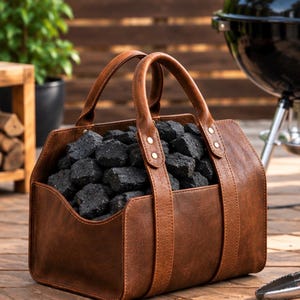 May include: A brown rectangular carrier with two sturdy handles and metal rivets, filled with black charcoal briquettes. The carrier features a smooth, leather-like texture and an open-top design, providing a convenient way to transport fuel.
