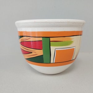 Può includere: Una ciotola in ceramica bianca con un bordo largo e una base arrotondata. La ciotola è decorata con un motivo geometrico di forme arancioni, rosse, verdi e beige. Il bordo presenta piccole macchie marroni.