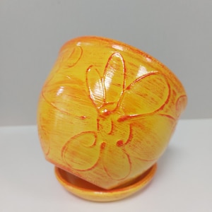 May include: A yellow ceramic planter with a raised orange floral design. The planter has a matching saucer.