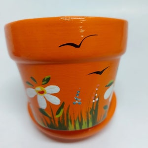 May include: Orange terracotta flower pot with a hand-painted design of white flowers, green grass, and black birds.