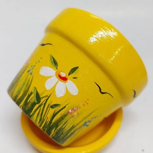 Puede incluir: Una maceta de cerámica amarilla con un diseño pintado de una margarita blanca y hierba verde. La maceta tiene un platillo amarillo a juego.
