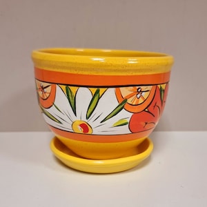 Puede incluir: Una maceta de cerámica pintada a mano con un platillo a juego. La maceta es amarilla con diseños florales naranjas y blancos. El borde y el platillo son de color amarillo liso. La maceta tiene unos 15 cm de diámetro.