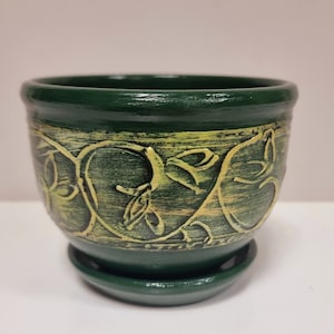 Op de afbeelding: Een donkergroene keramische plantenpot met een bijpassende schotel. De pot heeft een verhoogd, goudgeel bloemmotief rond het midden. De pot heeft een diameter van ongeveer 13 cm en is 10 cm hoog, ideaal voor kleine planten.