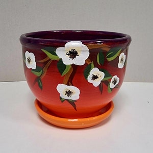Puede incluir: Una maceta de cerámica pintada a mano con un platillo a juego. La maceta es un degradado de rojo a naranja, con un borde morado. Flores blancas con centros negros y hojas verdes están pintadas en la maceta.