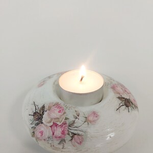 May include: A white, oval candle holder with a lit tea light. The holder features pink rose floral patterns. The candle holder is a decorative item.