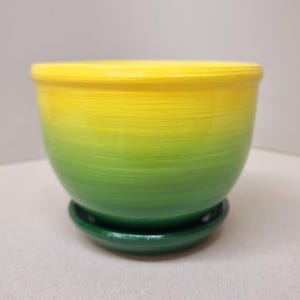 Könnte beinhalten: Ein runder Keramik-Pflanztopf mit Untersetzer in Ombre-Optik. Der Topf ist grün und gelb und hat eine glatte, glänzende Oberfläche.